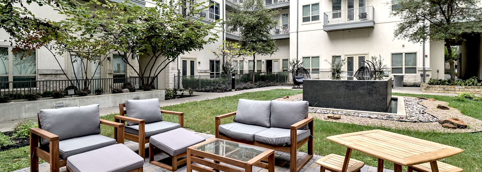 Looking down at grey cushioned outdoor furniture and a wooden table set sitting before the black, square fountain in the Zen Courtyard. The apartment building is seen behind.