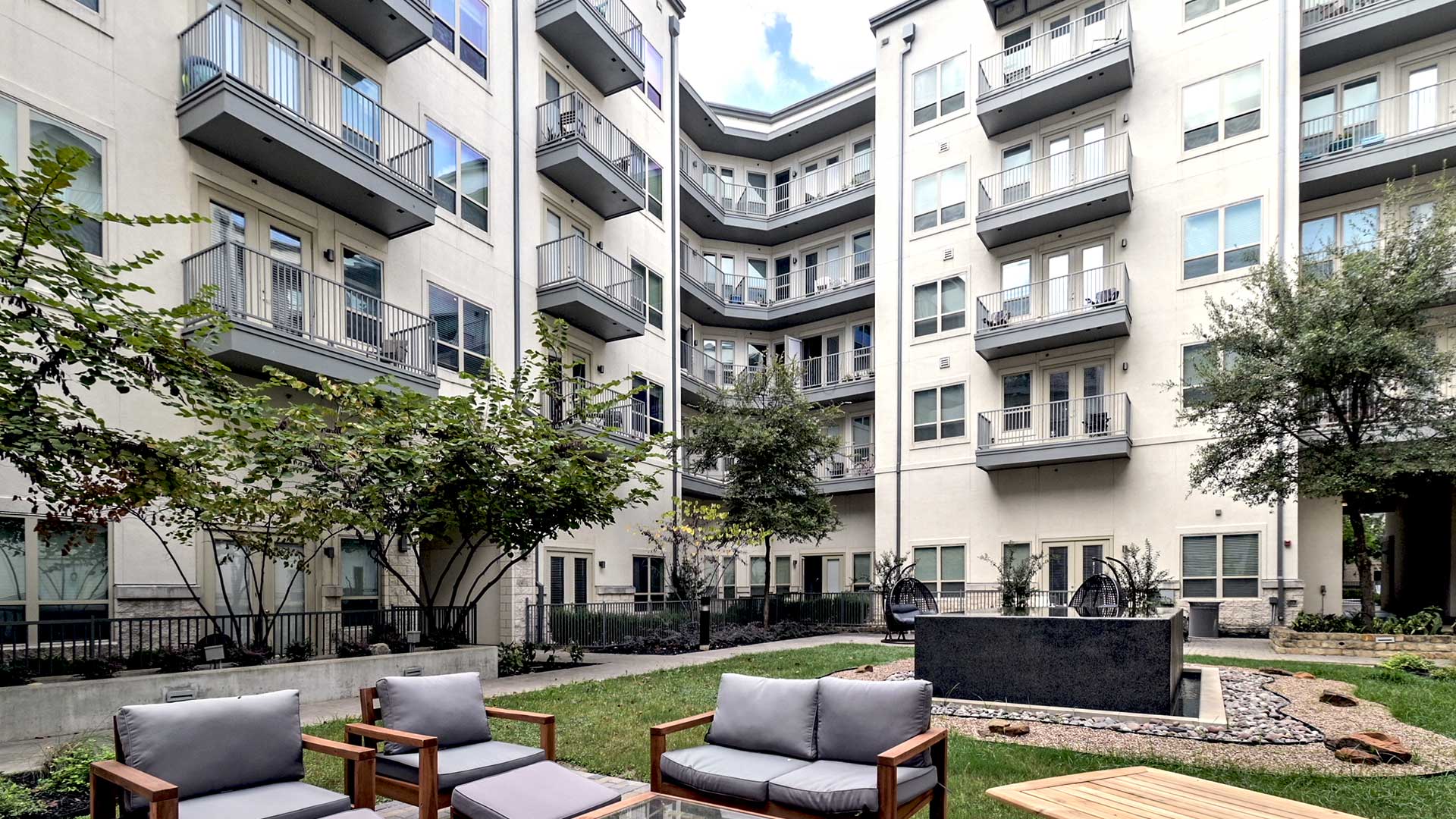 Looking at grey cushioned outdoor furniture and a wooden table set sitting before the black, square fountain in the Zen Courtyard. The apartment building is seen behind.