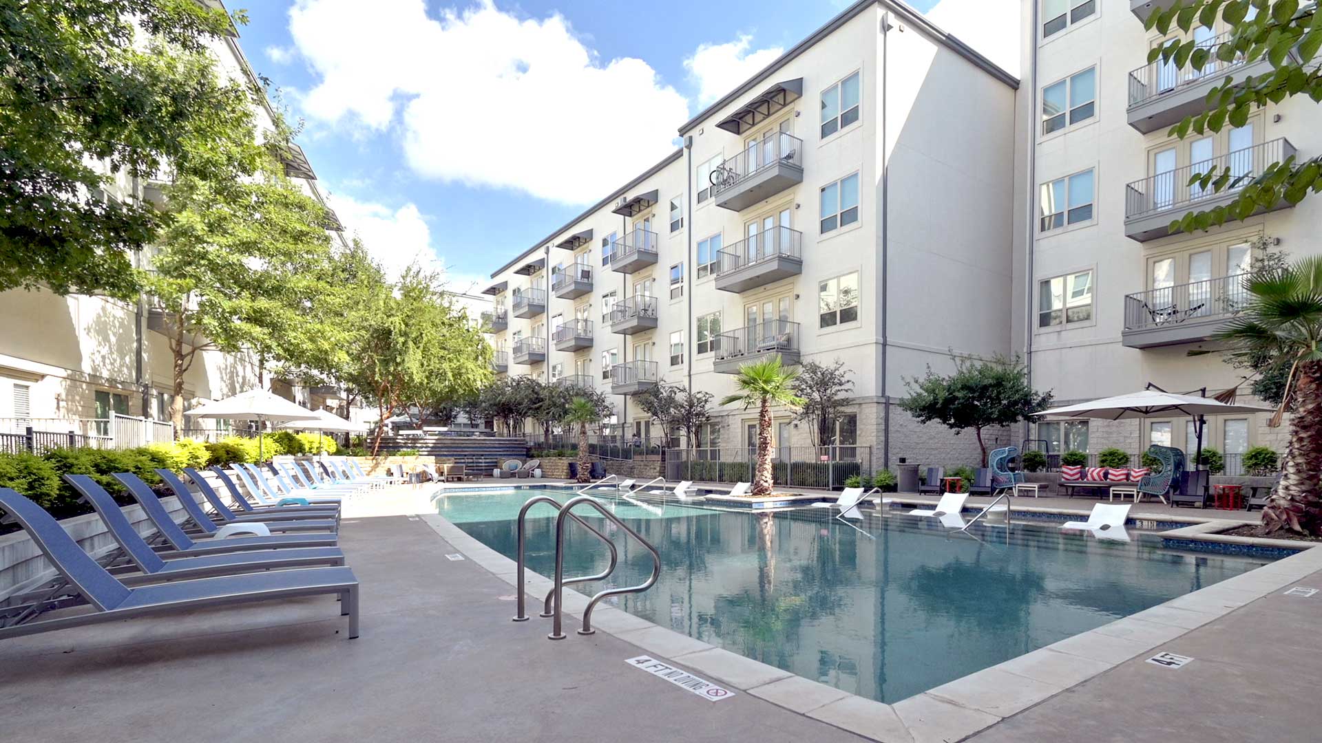 Standing at the corner of the pool with a line of lounge chairs running off down the left and the tanning shelf across the pool. There are trees all over the courtyard and the apartment building surrounds the scene.