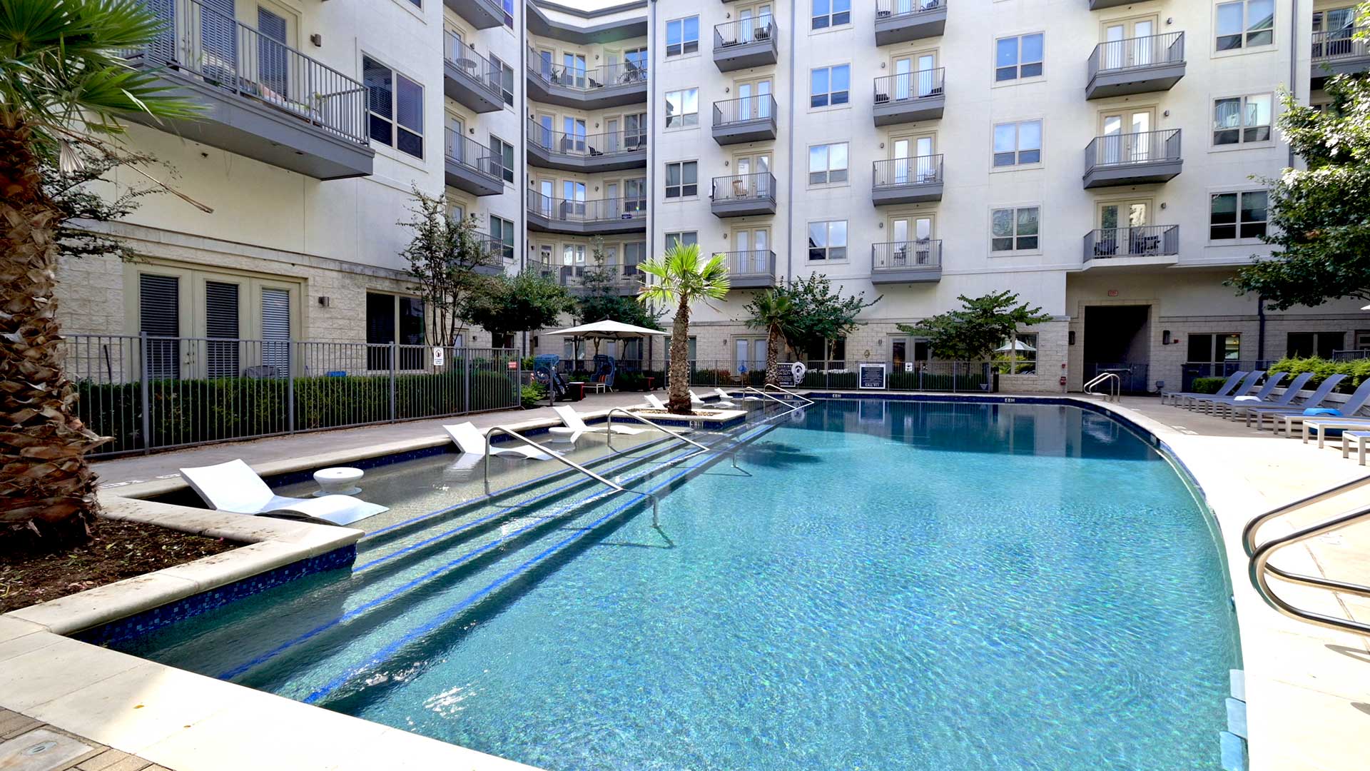 Looking down the outdoor pool with the apartment building at the far end. The tanning shelf runs along the left with a palm tree in the middle.