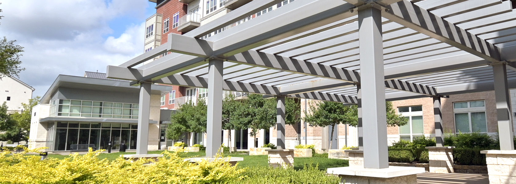 A large grey pergola with a patio beneath is before us. A grass field goes off to the left with the glass windows of the office building to Crest at Las Colinas Station at the end. The apartment building runs along the back.