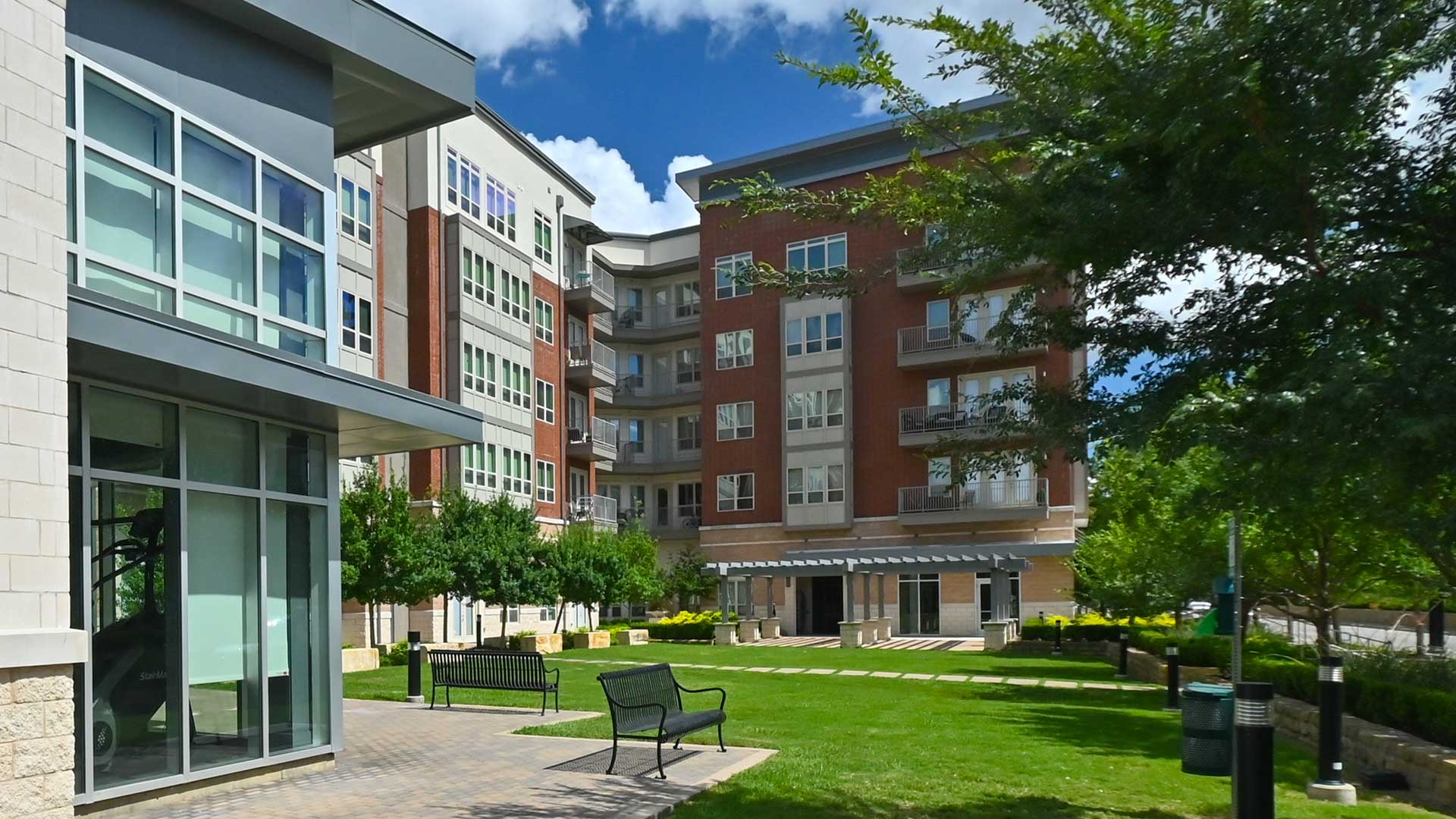 Looking across an open green space with the apartment building along the left and far side. Windows to the fitness center and a set of park benches on are to the left and trees line the right side.