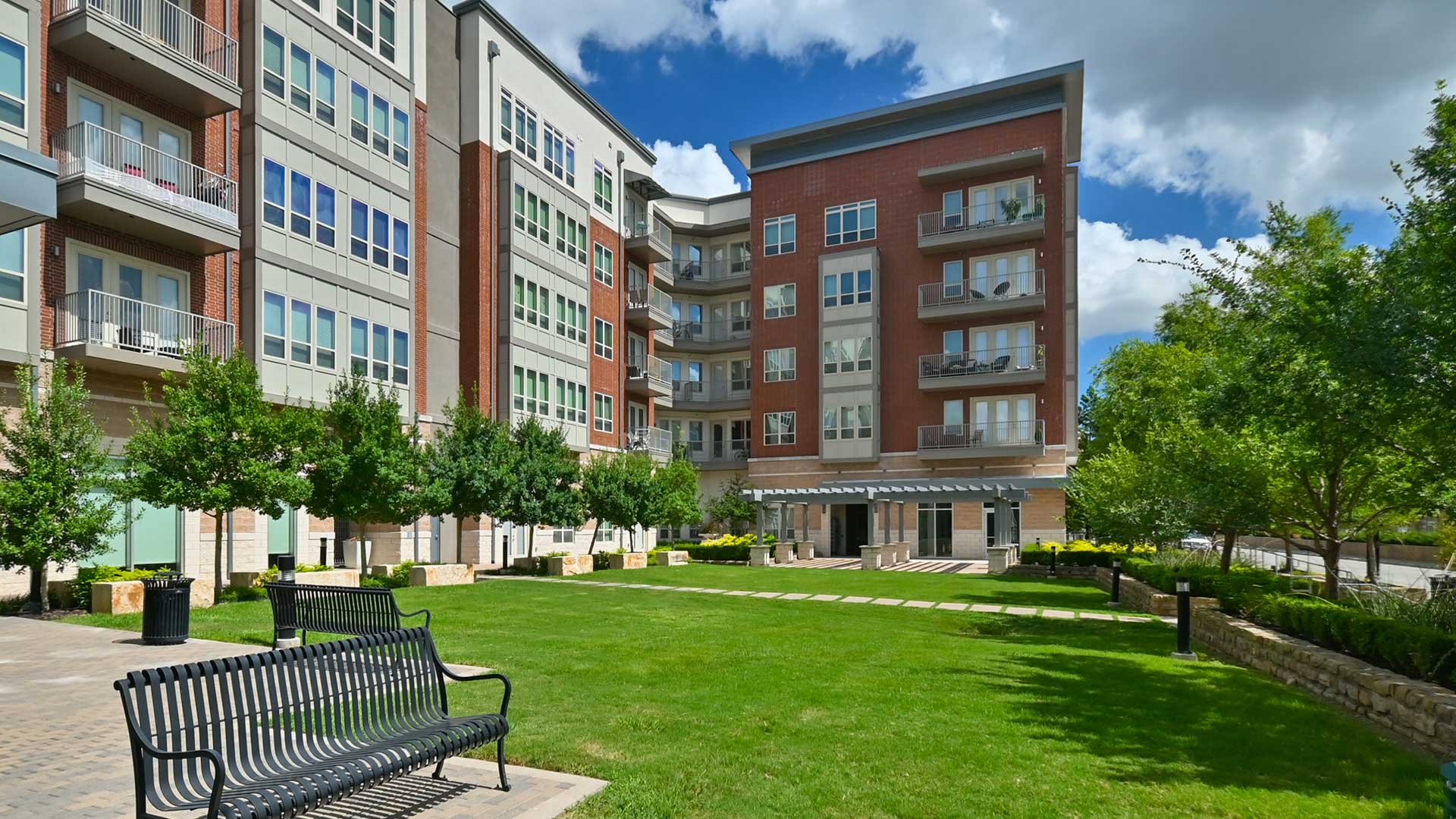 Looking across an open green space with the apartment building along the left and far side. A set of park benches on are to the left and trees line the right side.