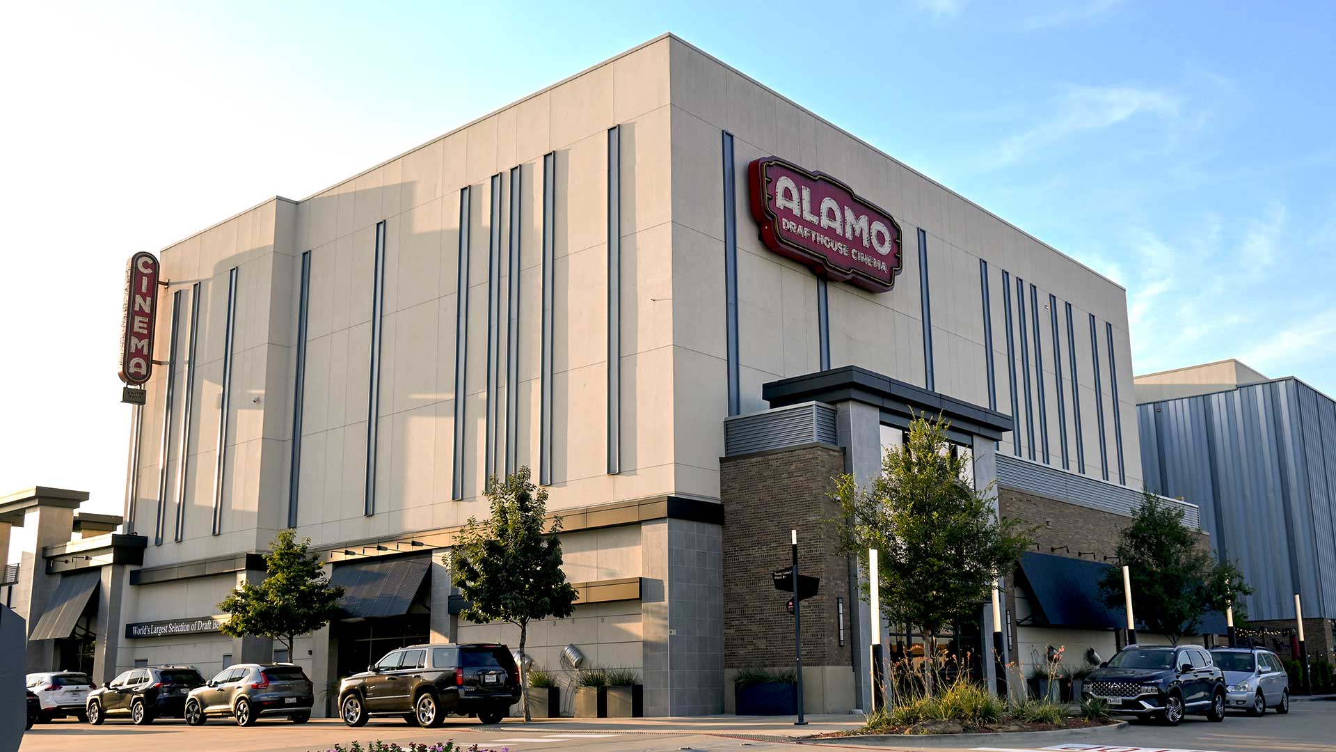 Looking at the square concrete building of the Alamo Drafthouse Cinema from across the street. There are cars parked in front of the building.