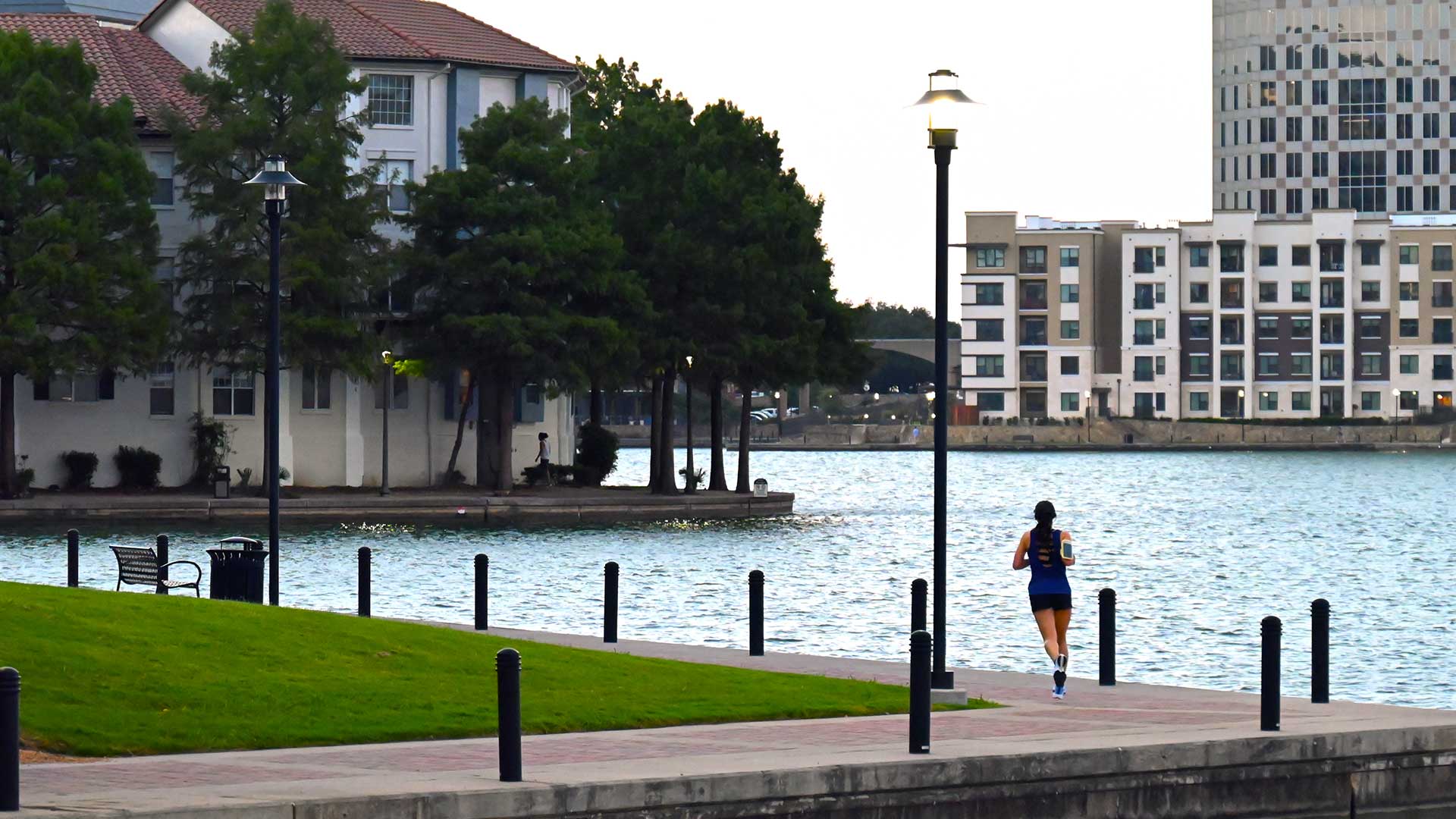 A woman jogs along a brick path along the shore of Lake Carolyn. The path heads off to the left where there are trees and a building. Another large building is seen across the lake on the right.
