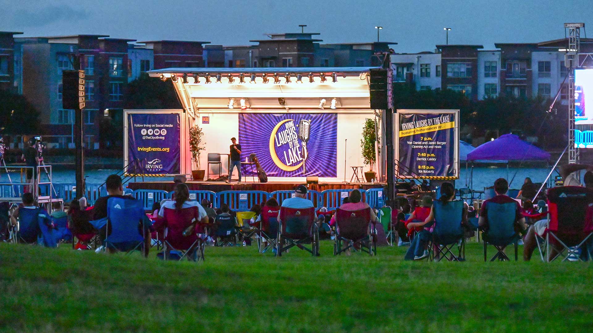 A stage is setup in front of a lake at night. People sit on a lawn in camping chairs watching a comedian on stage.