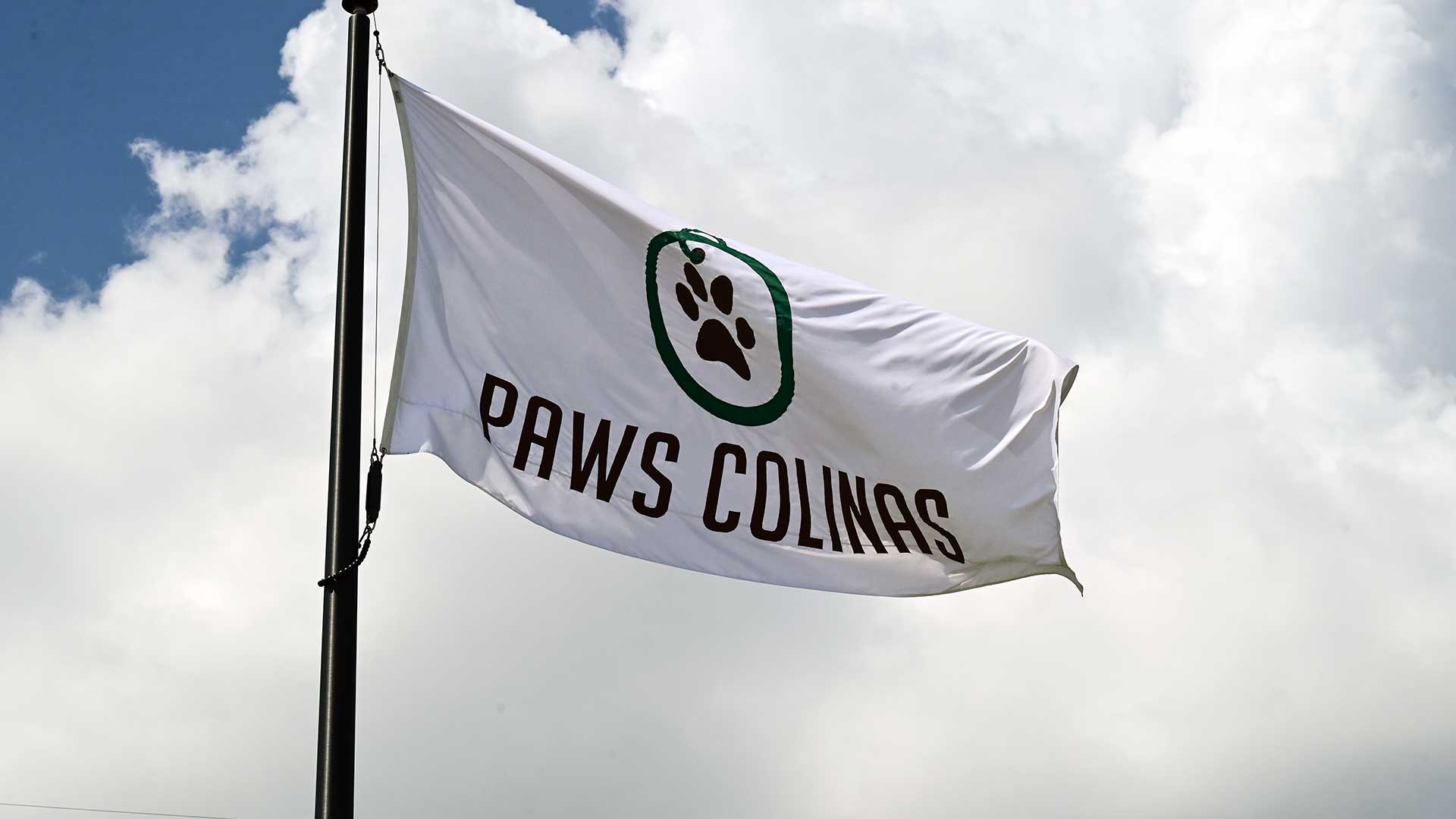 A close-up of the white flag that says "Paws Colinas" on it atop a flag pole on a cloudy day.