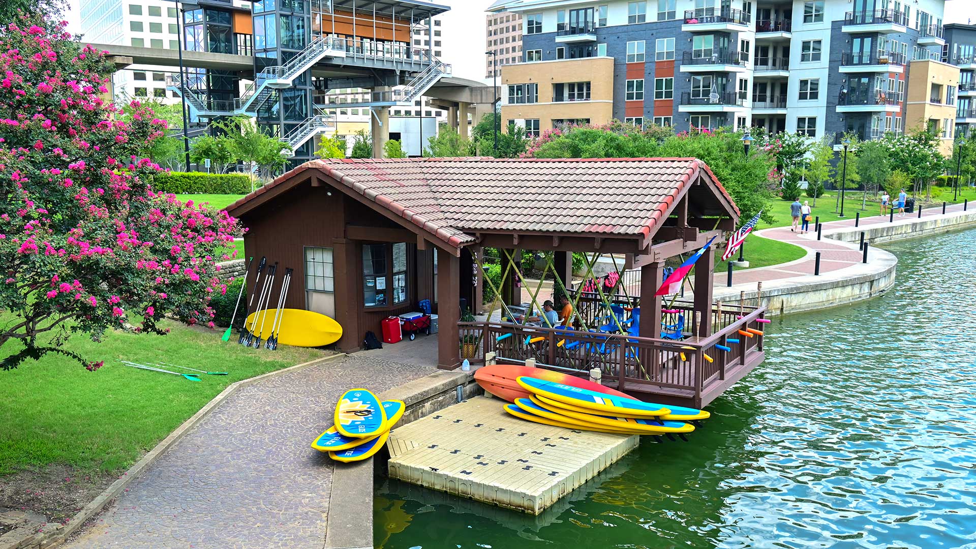 Looking down at a lakeside kiosk to rent kayaks and boards. There is a swim platform floating along the shore with several boards stack. A brick walkway follows the shoreline off to the right with apartment buildings behind.
