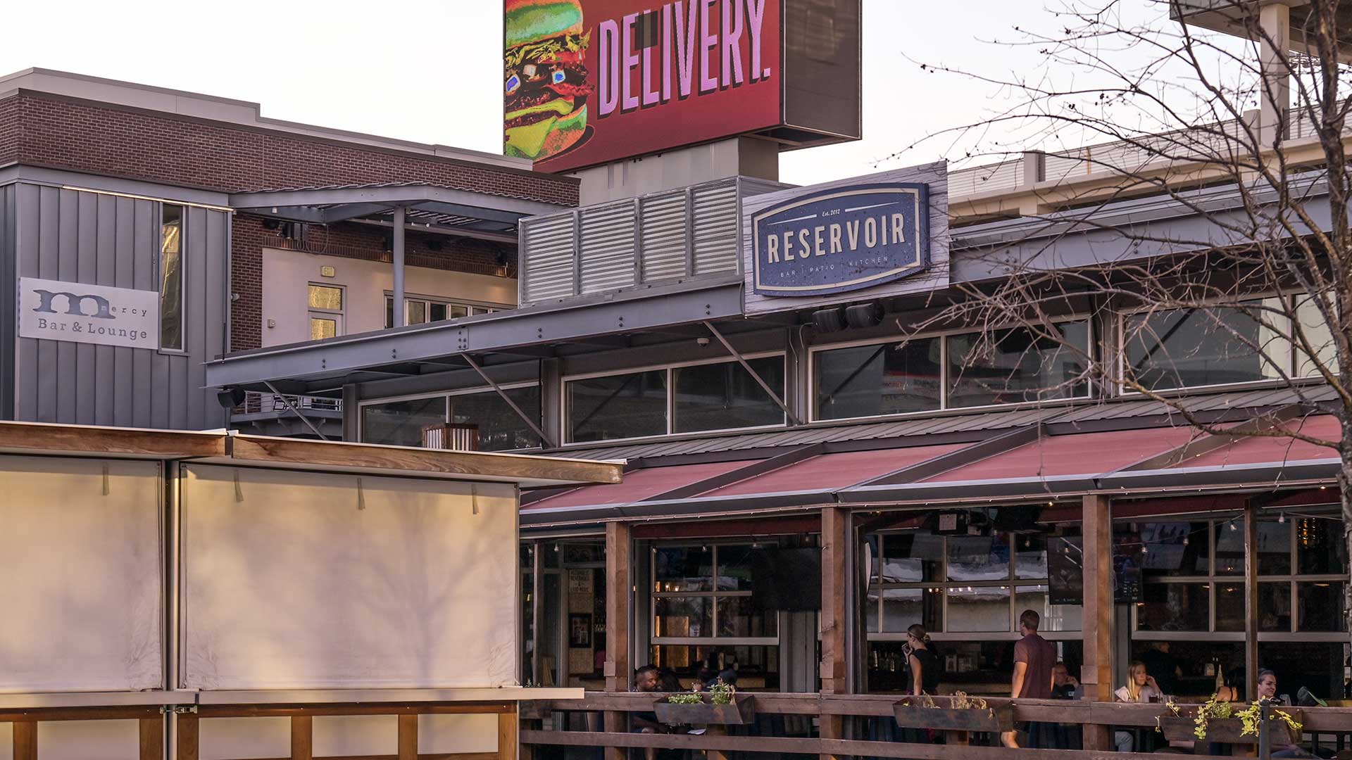 A close look at various outdoor patios at restaurants close to one another. There are signs above, including a large electronic screen.
