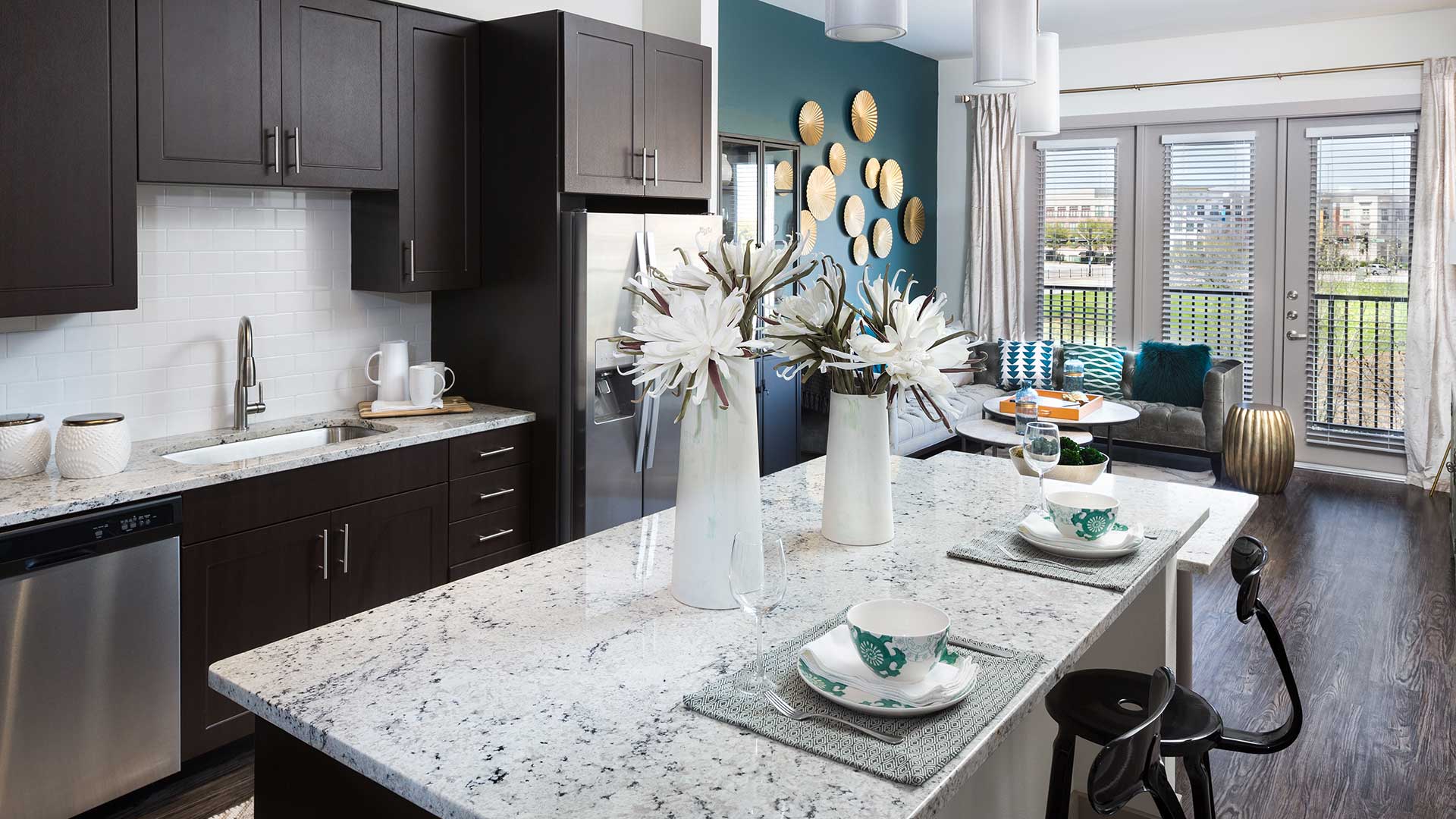 Looking along the kitchen island in a residence at Crest at Las Colinas Station. The kitchen runs along the left wall with ebony cabinets and stainless steel appliances. The island has two large flower vases and two place settings. There are two stools in front of the island on the right with the living room seen further behind.