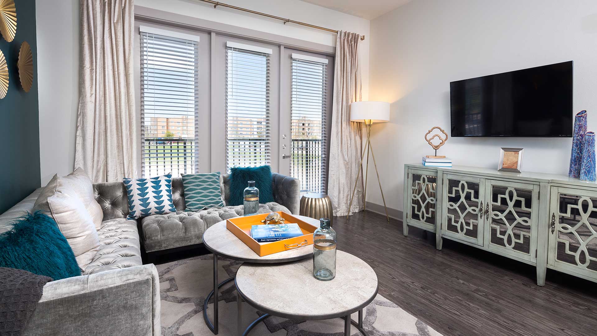 A living room in a residence at Crest at Las Colinas Station. A sectional couch is on the left with a set of round coffee tables before it. On the right is a dresser with various art pieces on it and a flat screen on the wall above. There are three large windows in the middle.