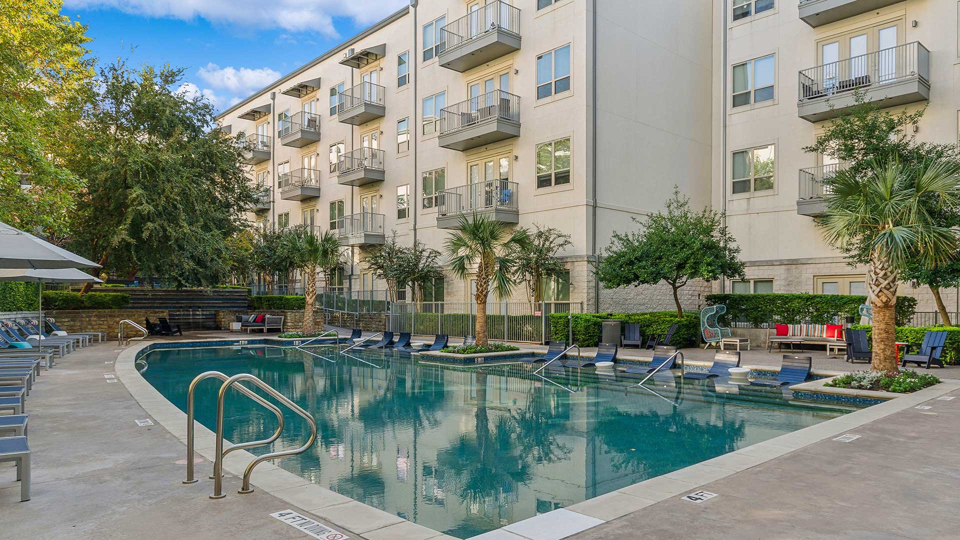 A serene outdoor pool area at an upscale residential community, surrounded by lush landscaping and tall palm trees. The pool features a set of in-water lounge chairs and a clean, paved pool deck with additional seating, including lounge chairs and shaded areas. In the background, a modern four-story apartment building with private balconies overlooks the pool. Cozy seating areas with colorful cushions enhance the inviting atmosphere.