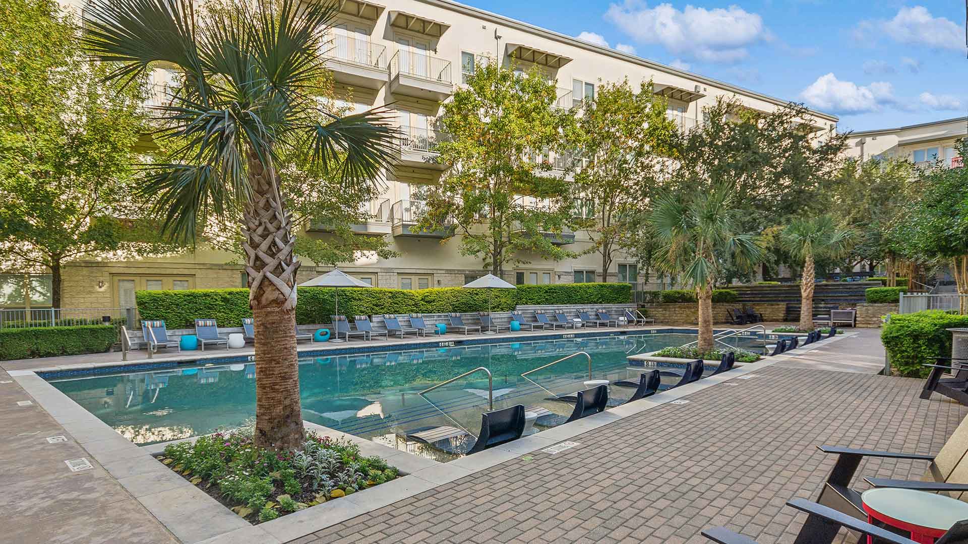 A vibrant outdoor pool area in a residential complex, featuring built-in water loungers and a paved pool deck lined with cushioned lounge chairs and shaded umbrellas. Lush greenery, including palm trees and well-maintained hedges, frames the pool, creating a serene atmosphere. The surrounding apartment building showcases private balconies with views of the pool. The seating area along the brick pathway includes modern chairs and small tables, enhancing the outdoor relaxation space.