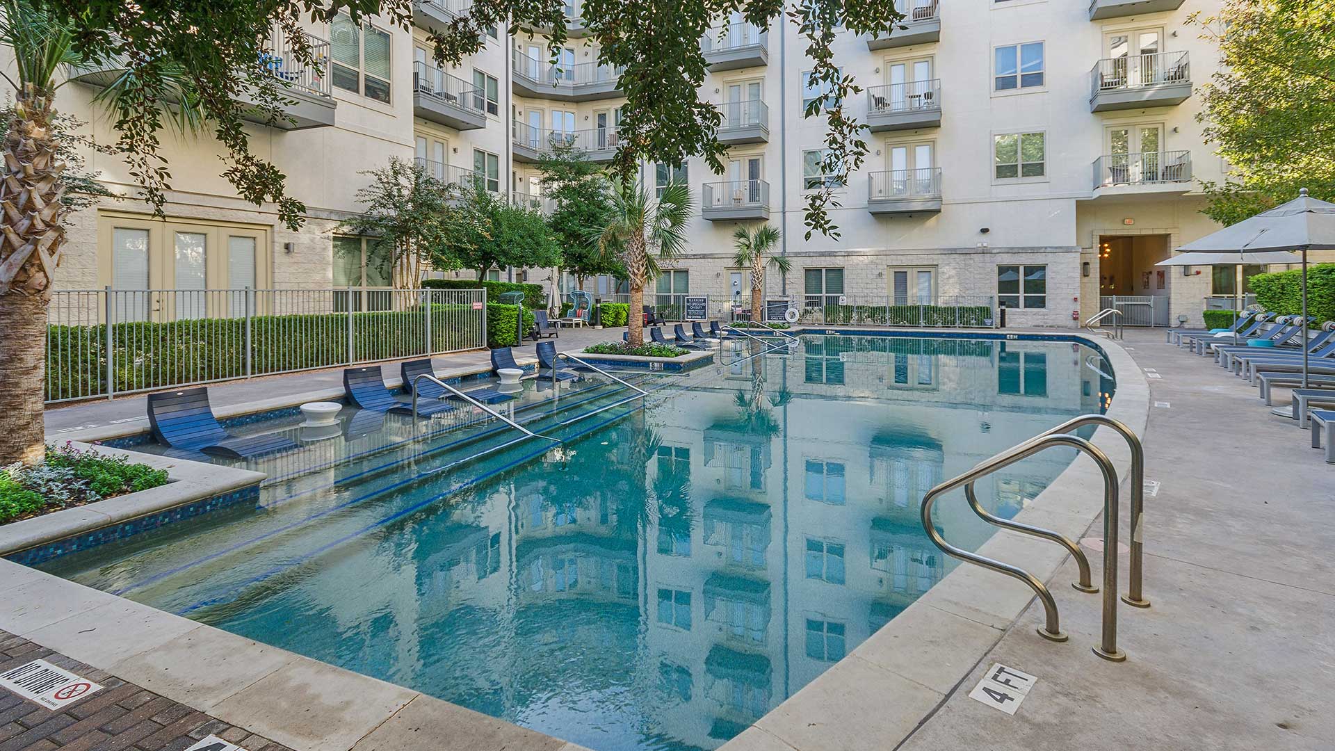 A tranquil outdoor pool area surrounded by a modern apartment complex. The pool features a series of built-in lounge chairs partially submerged in the water, offering a relaxing experience. The pool deck includes additional seating with cushioned lounge chairs and umbrellas for shade. The building’s balconies and windows reflect on the pool’s clear surface, while greenery and palm trees enhance the serene ambiance. A gated area with neatly trimmed hedges provides privacy and a touch of elegance.