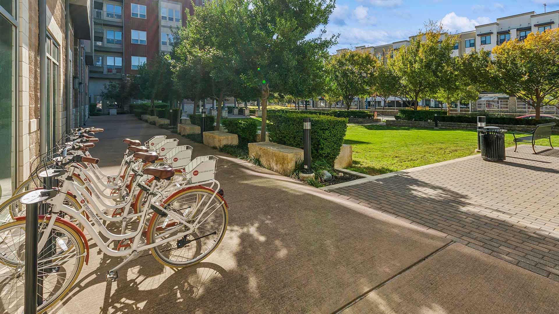 A sunny outdoor space at a residential community featuring a row of white bicycles with leather saddles and handlebars, available for residents. The bikes are parked along a paved pathway near a lush green lawn surrounded by neatly trimmed hedges and trees. Stone benches and a black metal trash can are placed along the path, offering convenience and seating. In the background, a modern building with large windows and balconies overlooks the landscaped area, creating a vibrant and inviting setting for relaxation or outdoor activities.