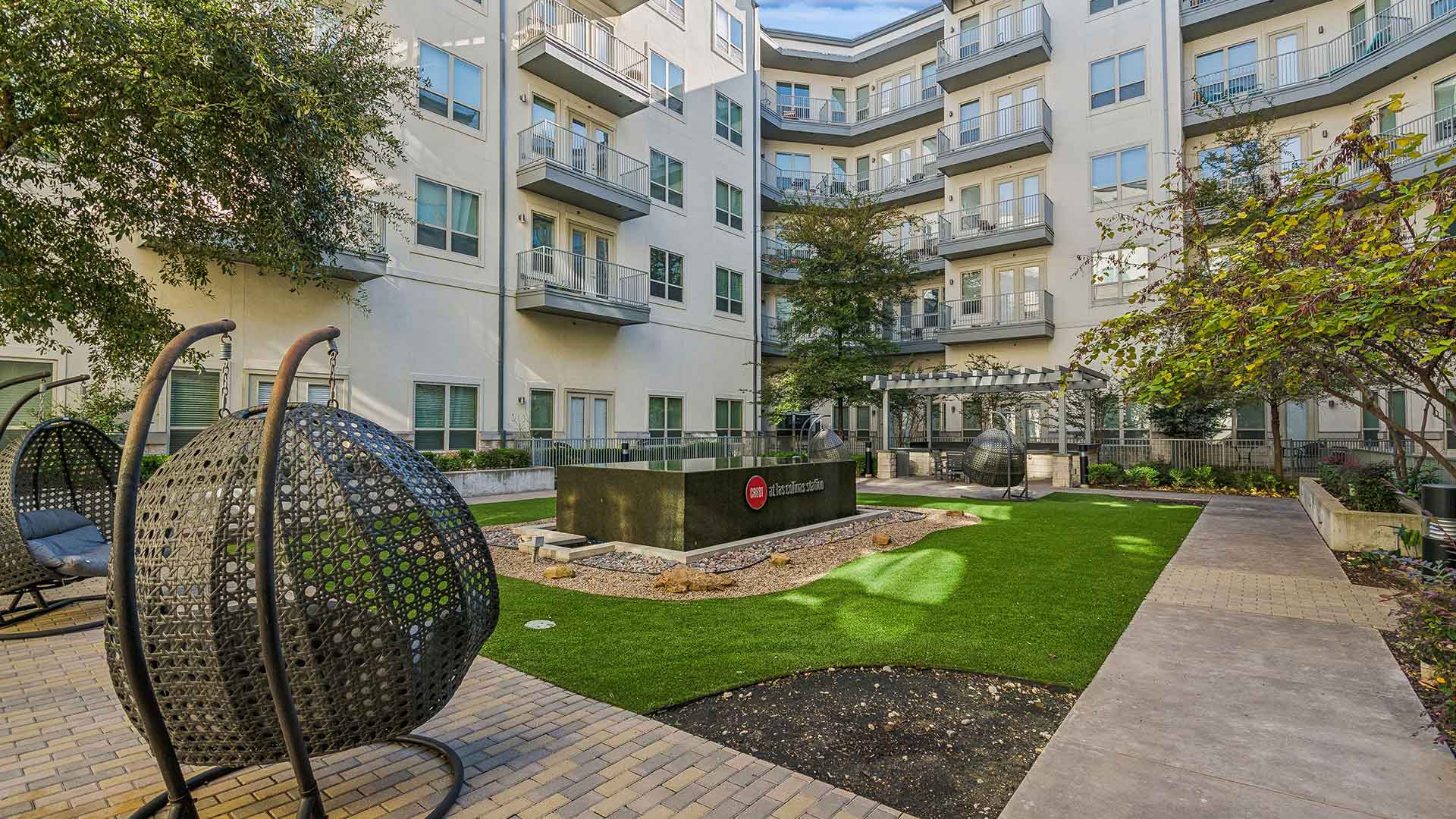 A peaceful courtyard in a residential complex featuring a modern water feature with the Crest Las Colinas logo, surrounded by decorative gravel and vibrant green artificial turf. The space includes unique woven hanging chairs with cushions, providing a relaxing seating option. A pergola with additional seating is visible in the background, surrounded by trees and well-maintained landscaping. The surrounding apartment building, with balconies overlooking the courtyard, enhances the serene and inviting atmosphere of this communal area.