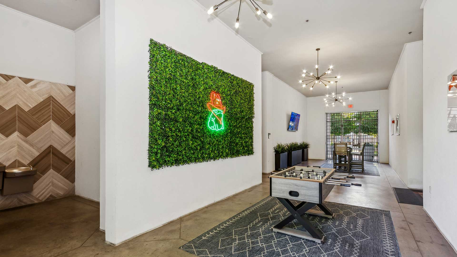 A stylish community lounge area featuring a modern foosball table on a geometric patterned rug. The space is decorated with a green living wall adorned with a neon sign of a cat wearing a cowboy hat, adding a playful touch. Contemporary lighting fixtures with starburst designs illuminate the room. In the background, there is a seating area with a large screen TV and a dining table. Potted greenery enhances the aesthetic, and a wall with chevron-patterned wood tiles adds texture and warmth to the design. The lounge opens to an outdoor view through glass doors, creating a bright and inviting atmosphere.