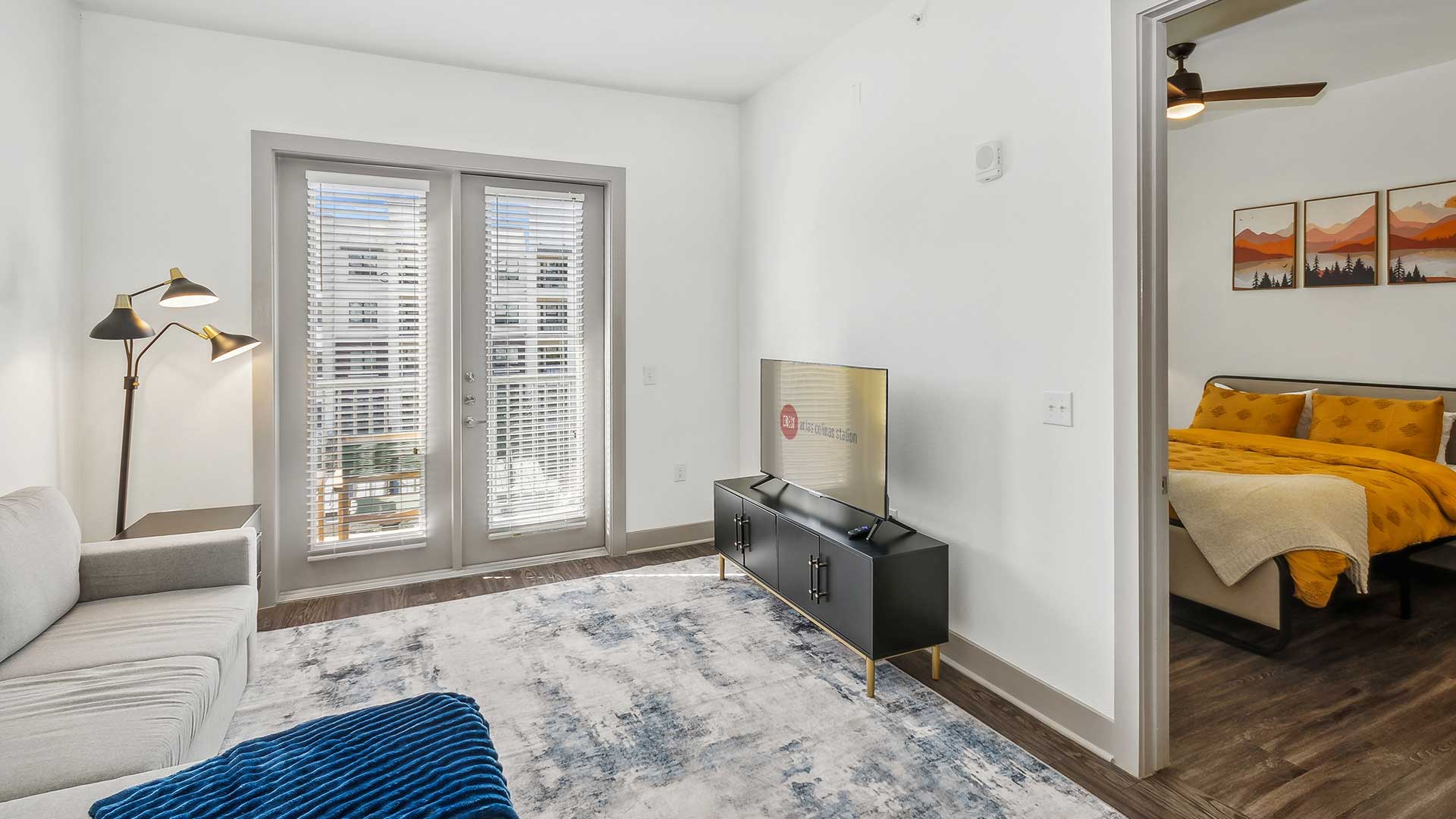 A cozy living room with a light gray sectional sofa, a blue textured throw blanket, and a soft area rug in shades of blue and white. The room features a floor lamp with three adjustable brass lights and a set of double glass doors leading to an outdoor space. A flat-screen TV sits on a modern black TV console.