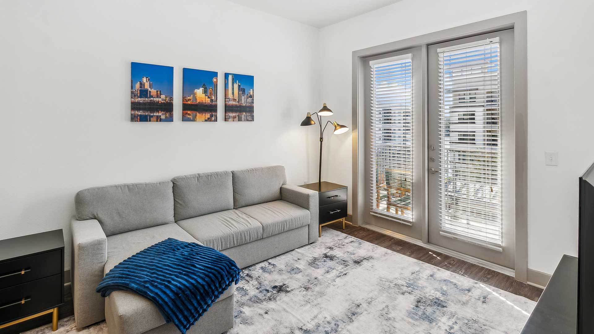 A similar angle of the living room with a focus on the sectional sofa, decorative blue throw, and nightstands on either side of the couch. Above the sofa are three framed prints showcasing a city skyline, adding color to the neutral walls.