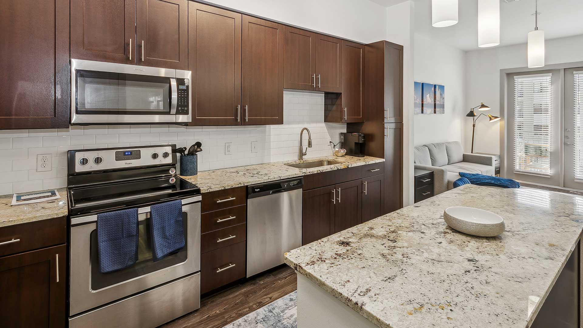 A contemporary kitchen with dark wood cabinetry, stainless steel appliances, and a granite countertop. The kitchen includes a modern electric range with a microwave above it, a dishwasher, and a subway tile backsplash.