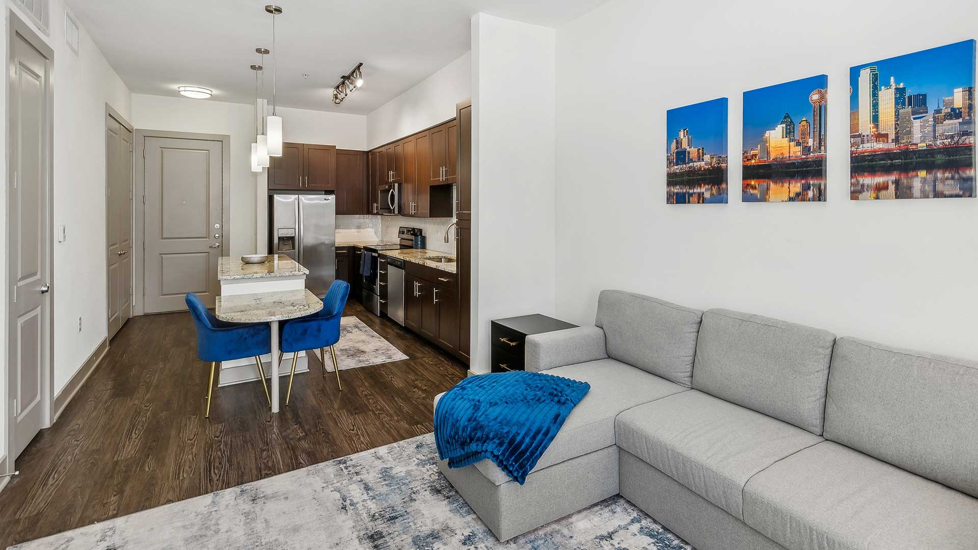 A wider angle of the kitchen and dining area, showing a small island with a granite surface and two blue upholstered dining chairs. The open-concept design flows into the living room, emphasizing the bright, airy layout.